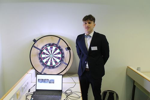 Ein junger Mann in einem Anzug steht neben einem Dartboard. Ein Laptop befindet sich auf einem Schreibtisch mit einer kabelgebundenen Verbindung.