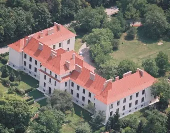 Ein großes weißes Gebäude mit einem roten Dach und mehreren Fenstern ist von Bäumen und Grünflächen umgeben. Das Gebäude hat mehrere Schornsteine und befindet sich in einer üppigen grünen Landschaft.