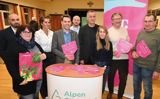 Eine Gruppe von Menschen steht hinter einem Tisch und hält pinke Schilder in der Hand. Sie lächeln und posieren für ein Foto. Auf dem Tisch steht das Wort 'Alpen'. Dahinter befindet sich ein großes Banner in Rosa und Weiß mit Text.