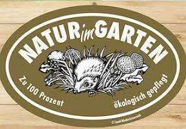 Ein braunes ovales Etikett mit 'Natur im Garten' in weiß, das einen Igel unter Pflanzen zeigt und 'Zu 100 Prozent' sowie 'ökologisch gepflegt' besagt. Auf einer Holzoberfläche platziert.