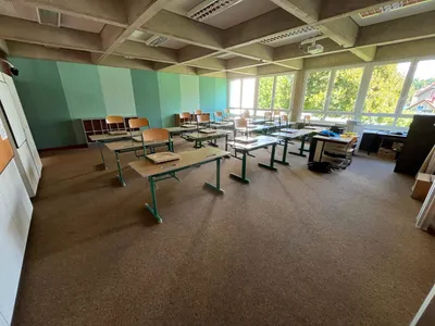 Ein leerer Klassenraum mit grünen und weißen Wänden, großen Fenstern und Reihen von Schreibtischen und Stühlen. Der Raum hat einen Teppichboden und einen Projektor an der Decke.