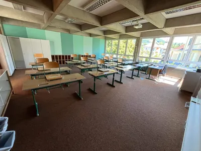 Ein leeres Klassenzimmer mit grünen Tischen und Stühlen, Teppichboden, grün-weißen Wänden und Glasfenstern mit Blick auf Bäume und ein Gebäude.