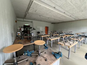 Ein Werkstattraum mit Holztischen und Metallstühlen. Geräte wie Bohrer und Zwingen sind auf den Tischen platziert. Eine Tafel ist an der Wand montiert. Der Raum ist mit Leuchtstofflampen gut beleuchtet.