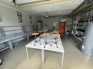 Ein leerer Raum mit weißen Wänden und Tischen mit Holzsitzen. Es gibt elektrische Schalttafeln an der Wand und Rohre an der Decke.