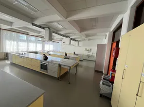 Eine leere, saubere Küche mit Edelstahlarbeitsplatten, gelben Schränken, Öfen und einer Spüle. Große Fenster und ein Feuerlöscher sind sichtbar.