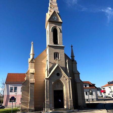 Eine Steinkirche mit einem Kirchturm und einer Uhr darauf, unter einem klaren blauen Himmel. Die Kirche hat einen spitzen Eingang mit einem Buntglasfenster darüber.