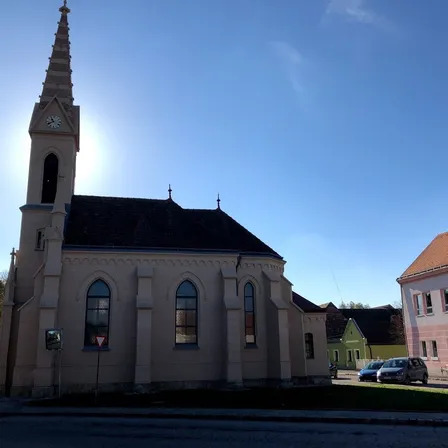 Eine Kirche mit Turm und Uhrenturm steht an einem sonnigen Tag. Ein rotes Schild ist davor zu sehen. Ein Auto ist am Straßenrand geparkt.