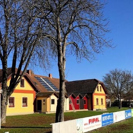 Zwei Häuser in einem ländlichen Gebiet mit Solarpanelen auf dem Dach. Die Häuser sind gelb und rot. Vor den Häusern stehen Bäume und ein Fußballfeld.