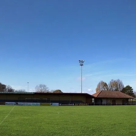 Ein großes Grasfeld unter einem klaren blauen Himmel. In der Mitte stehen zwei Laternenmasten und ein gelbes Haus in der Ferne.