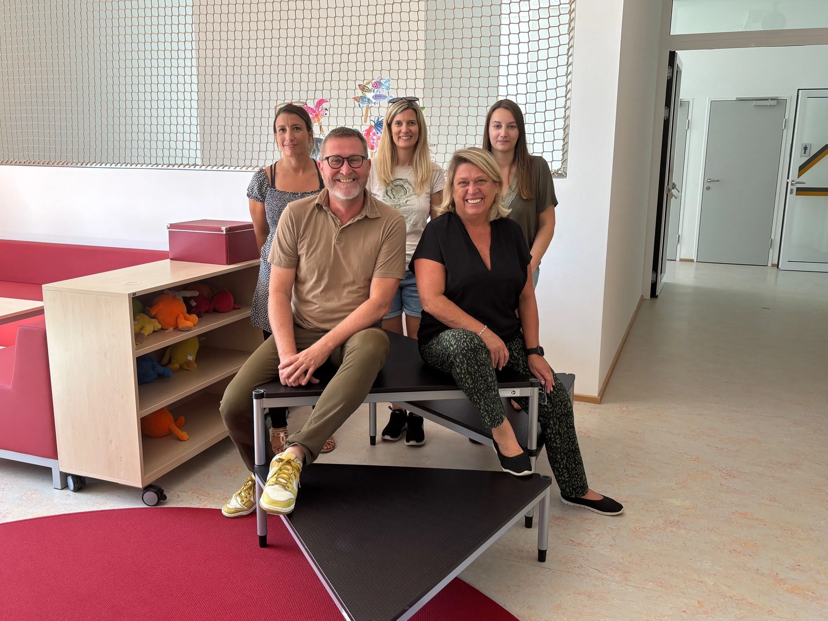 Fünf Erwachsene, drei Frauen und zwei Männer, posieren für ein Foto in einem Raum mit einer Wand, die ein Netz und bunte Designs aufweist. Sie sitzen auf einer Bank mit einem roten Teppich auf dem Boden.