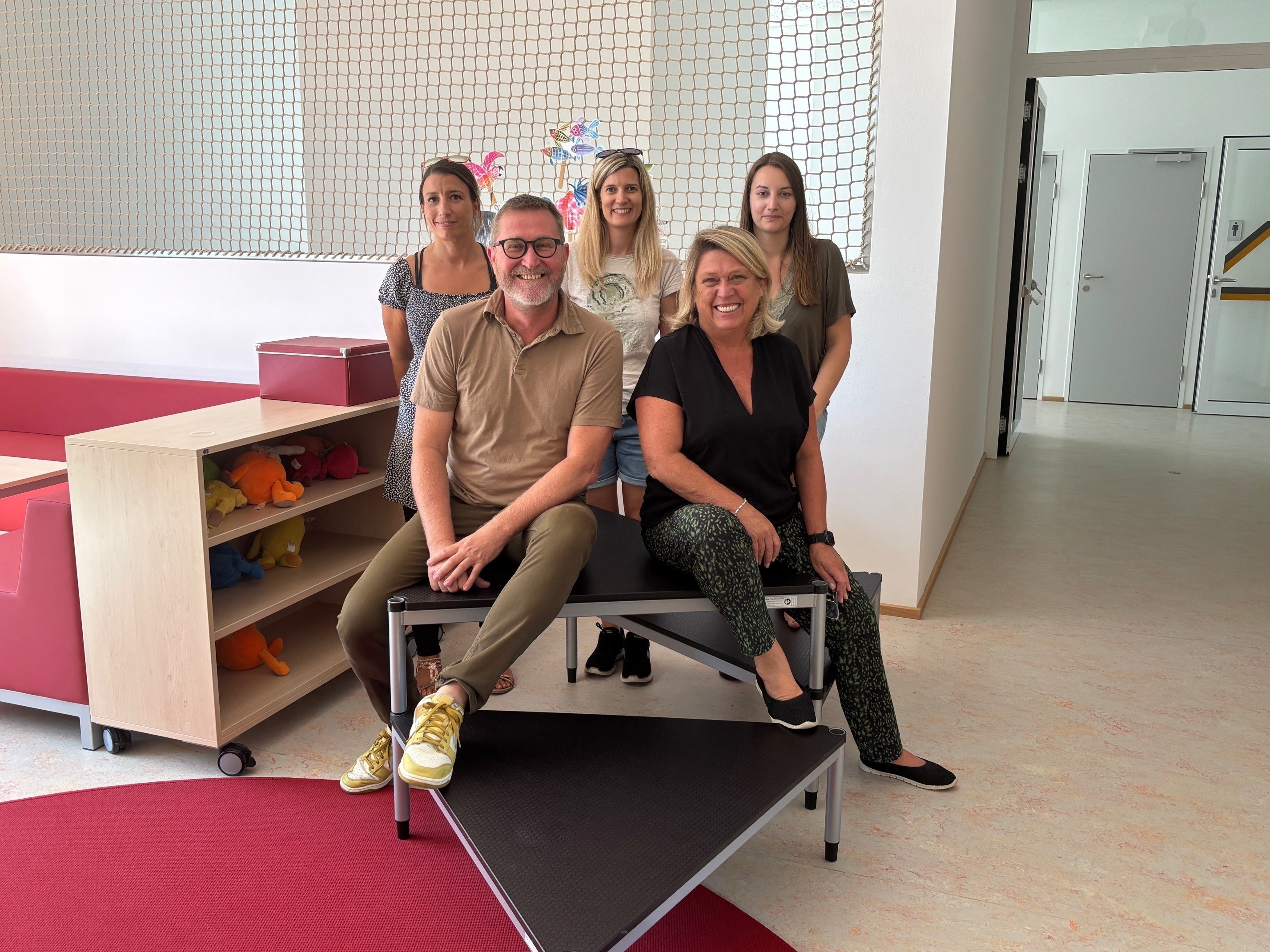Fünf Erwachsene, drei Frauen und zwei Männer, posieren für ein Foto in einem Raum mit einer Wand, die ein Netz und bunte Designs aufweist. Sie sitzen auf einer Bank mit einem roten Teppich auf dem Boden.