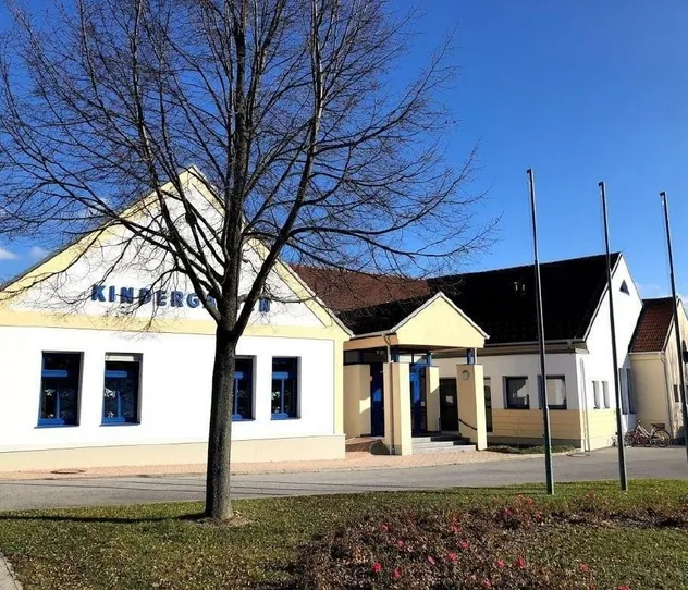 Ein Gebäude mit dem Wort Kindergarden an der Wand hat mehrere Fenster, eine Tür und zwei Fahnenmasten. Ein Baum steht vor dem Gebäude.