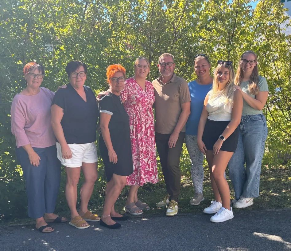 Eine Gruppe von Menschen, sowohl Männer als auch Frauen, posiert für ein Foto im Freien. Sie stehen vor einigen Bäumen und tragen Freizeitkleidung. Sie alle lächeln und schauen in die Kamera.
