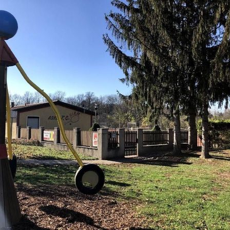 Eine Außenszene mit einer einzigartigen Kunstinstallation, die einem Besen ähnelt. Sie befindet sich in einem Park, mit einem Gebäude und einem Basketballkorb im Hintergrund.
