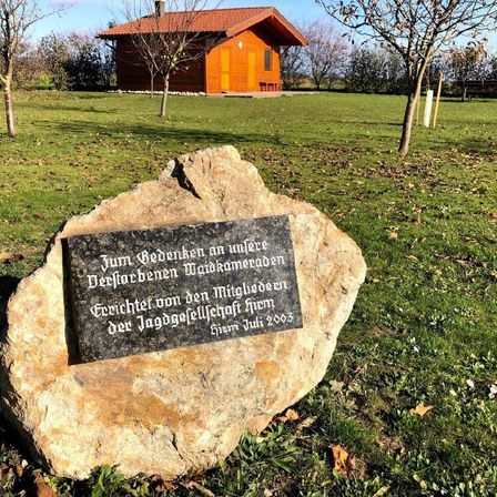 Ein Stein mit einer Plakette in einem Grasfeld, mit einem kleinen Holzhaus und Bäumen im Hintergrund.