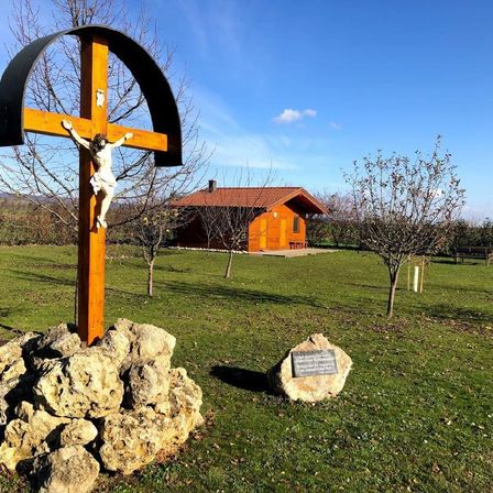 Ein hölzernes Kreuz mit einem Kruzifix in einem Feld. Im Hintergrund befinden sich ein Haus und eine Bank. Das Feld ist von Bäumen umgeben.