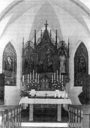 Ein altes Foto des Inneren einer Kirche mit einem Altar vorne. Der Altar hat ein Kreuz, Statuen und Kerzen. Es gibt Buntglasfenster auf beiden Seiten.