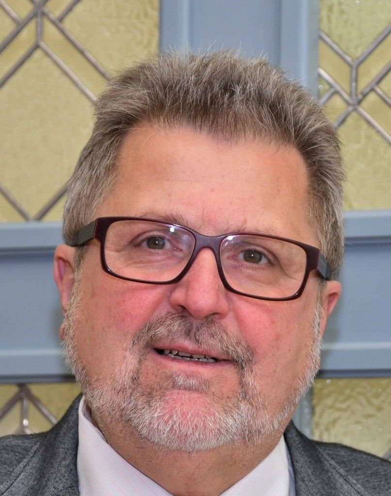 Ein Nahaufnahme des Gesichts eines Mannes, der eine Brille, ein weißes Hemd und einen grauen Anzug trägt. Er hat einen grauen Bart und Schnurrbart und ein leichtes Lächeln auf dem Gesicht. Der Hintergrund zeigt eine gelbe Wand mit einem Diamantmuster.