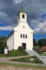 Eine kleine weiße Kirche mit einem grünen Turm und einem Kreuz auf der Spitze. Die Kirche ist von Gras und Pflanzen umgeben.