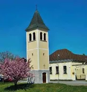 Eine Kirche mit einem hohen Turm und einem Kreuz auf der Spitze, umgeben von einem Rasenbereich und einem rosa blühenden Baum.