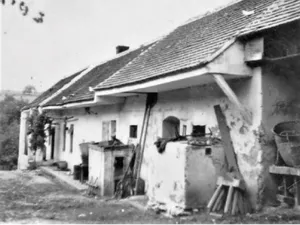 Ein Schwarz-Weiß-Foto eines alten Hauses mit einem Ziegeldach und einem Schornstein. Draußen befinden sich mehrere Gegenstände, darunter ein Fass, eine Leiter und ein Holzstapel.