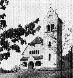 Ein Schwarz-Weiß-Foto einer Kirche mit Turm und Kreuz oben, umgeben von Bäumen und auf einem Hügel gelegen.