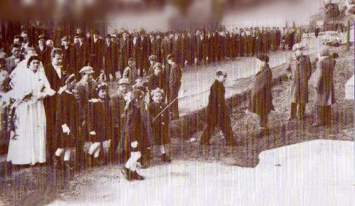 Ein Schwarz-Weiß-Foto zeigt eine Menschenmenge auf einer Straße, vermutlich für eine Zeremonie, einige tragen Hüte und halten Blumen.