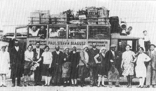 Ein Schwarz-Weiß-Foto einer Gruppe von Menschen, die vor einem Bus mit der Aufschrift 'Paul Stefan Blagusz' stehen. Sie sind in Vintage-Kleidung gekleidet.