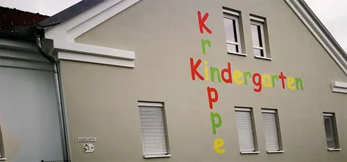Ein Gebäude mit drei Fenstern hat ein Schild mit der Aufschrift 'Kr Kindergarten Pe'. Das Gebäude hat eine weiße Außenseite und ein graues Dach.