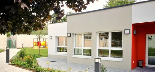 Ein modernes, beigefarbenes Gebäude mit weißem Fensterrahmen und einem Spielplatz im Hintergrund.