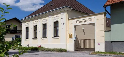 Ein Kindergartengebäude mit einem braunen Dach, cremefarbenen Wänden und mehreren Fenstern. Über dem Eingang befindet sich ein Schild mit der Aufschrift 'Kindergarten'.