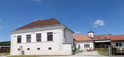 Ein weißes Gebäude mit braunem Dach und mehreren Fenstern steht draußen. Es hat ein Schild, auf dem 'Volksschule' steht. In der Nähe sitzen Kinder auf Stühlen an einem Tisch im Freien.
