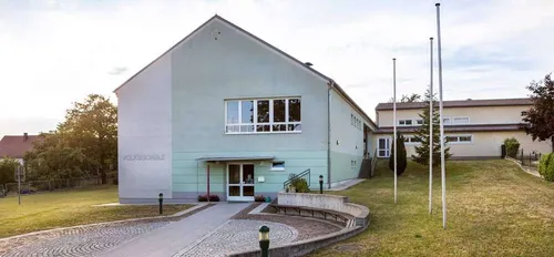 Das Volksschule-Gebäude hat ein modernes architektonisches Design mit Glasfenstern und einem Weg, der zum Eingang führt. Der Eingang hat eine Glastür und ein Metallgeländer. Das Gebäude ist von einem Grasbereich mit Pflanzen und Bäumen umgeben.