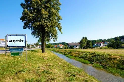 Ein Dorf namens Weppersdorf wird gezeigt, mit einem Schild auf der linken Seite, einem kleinen Fluss in der Mitte und Häusern in der Ferne.