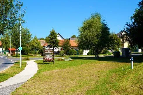 Ein Gemeinschaftspark mit einem gepflasterten Gehweg und einem Schild, umgeben von Häusern, Bäumen und einem klaren blauen Himmel.