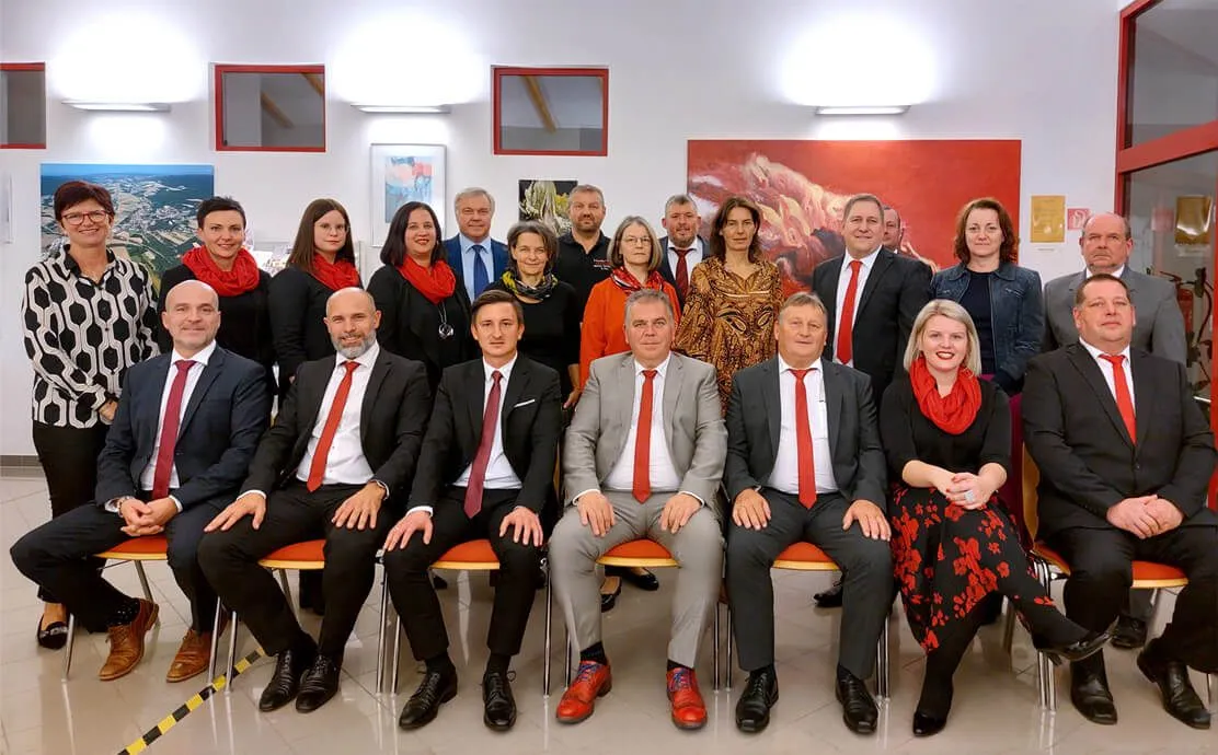 Eine Gruppe formell gekleideter Personen sitzt auf Stühlen in einem Besprechungsraum, mit einem großen Gemälde an der Wand hinter ihnen.