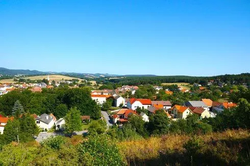 Ein kleines Dorf, eingebettet zwischen Bäumen und Hügeln unter einem klaren blauen Himmel. Die Häuser sind in verschiedenen Farben gestrichen, wobei orange Dächer hervorstechen.