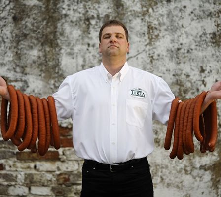 Ein Mann steht vor einer Backsteinmauer, trägt ein weißes Hemd mit 'TURZA' darauf gedruckt und hält mehrere Würste in beiden Händen.