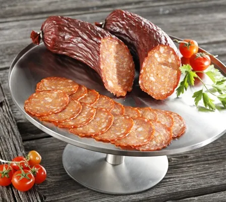 Ein Teller mit geschnittener Salami und ganzen Würsten auf einem silbernen Ständer, mit Kirschtomaten und Petersilie auf einer Holzoberfläche.