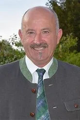 Ein Mann in einer grauen Weste und grüner Krawatte, mit einem Schnurrbart und einem Lächeln, steht vor einem unscharfen Hintergrund von Bäumen.