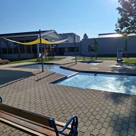 Freibad mit Sonnensegel und einer Bank. Im Hintergrund ein Gebäude mit einem gelben Rutschen und einem Baum.