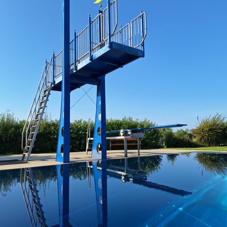 Ein blauer Sprungturm mit zwei gelben Fahnen steht über einem klaren Wasserbecken. Eine Leiter führt zur Plattform, und der Himmel ist klar und blau.