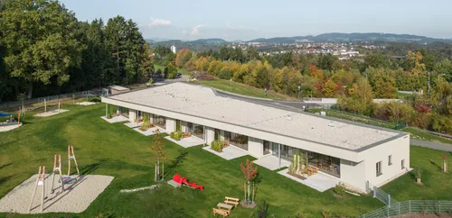 Luftaufnahme eines modernen Gebäudes mit Glaswänden, einem geschwungenen Dach und einem grünen Rasen mit Bänken und einer roten Skulptur.