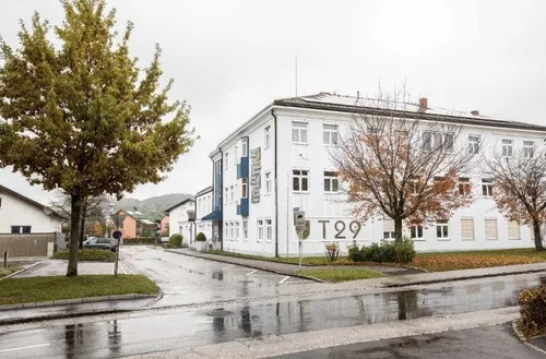 Ein weißes Gebäude mit vielen Fenstern und einem blauen Vordach befindet sich auf einer nassen Straße. Bäume, Büsche und ein geparktes Auto sind ebenfalls im Bild.