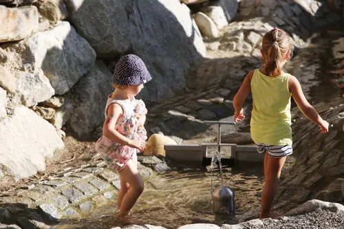 Zwei kleine Mädchen spielen in einer Wasserfontäne, umgeben von Felsen in einer sonnigen Außenumgebung.