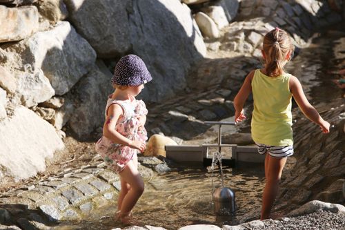Zwei kleine Mädchen spielen in einer Wasserfontäne, umgeben von Felsen in einer sonnigen Außenumgebung.