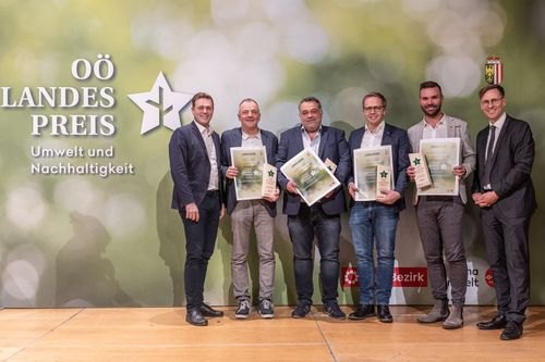 Fünf Männer stehen auf einer Bühne und halten Urkunden in den Händen. Sie lächeln und scheinen für ein Foto zu posieren. Der Hintergrund zeigt einen Stern und Text in deutscher Sprache.