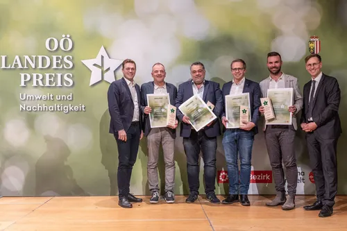 Fünf Männer stehen auf einer Bühne und halten Urkunden in den Händen. Sie lächeln und scheinen für ein Foto zu posieren. Der Hintergrund zeigt einen Stern und Text in deutscher Sprache.