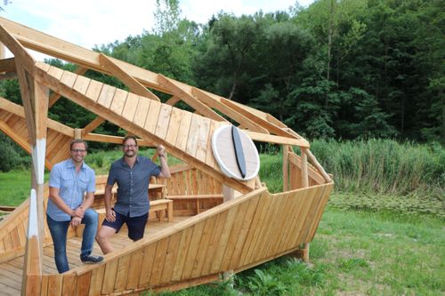 Zwei Männer in Freizeitkleidung posieren vor einer hölzernen Struktur, die einem Boot ähnelt, in einem üppigen grünen Wald.