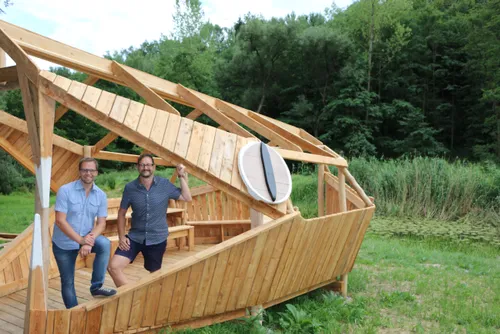 Zwei Männer in Freizeitkleidung posieren vor einer hölzernen Struktur, die einem Boot ähnelt, in einem üppigen grünen Wald.